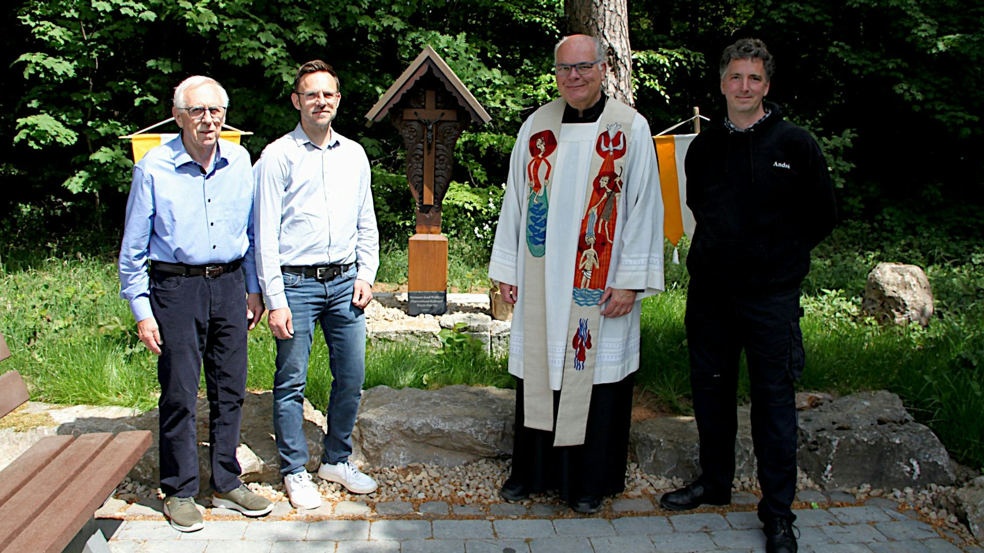 Das Bild zeigt vier Männer, die neben einer Sitzbank vor einem Wegekreuz stehen.
