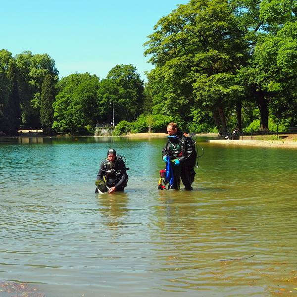 Zwei Männer in Taucheranzügen stehen in einem Weiher. Einer hat eine Pflanzkiste in der Hand.