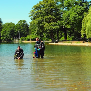 Zwei Männer in Taucheranzügen stehen in einem Weiher. Einer hat eine Pflanzkiste in der Hand.