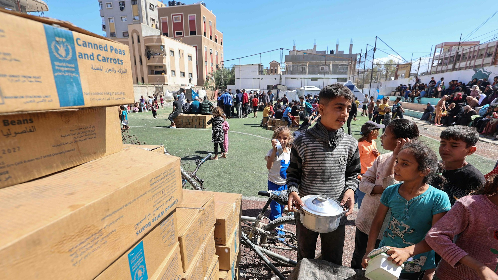Kinder mit Schüsseln stehen vor Pappkisten mit Essenslieferungen.