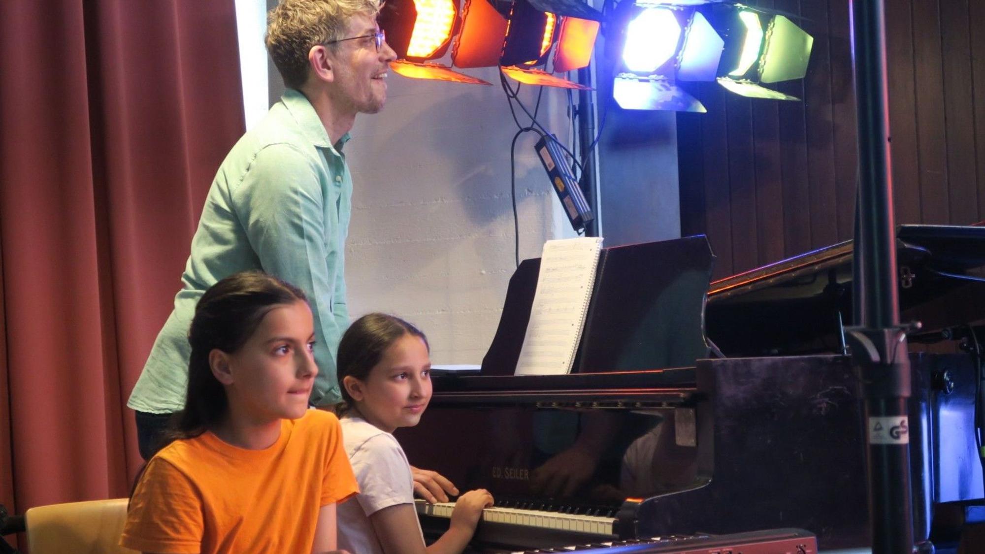 Ein Mann steht am Klavier, zwei Mädchen sitzen an Keyboard und Piano.