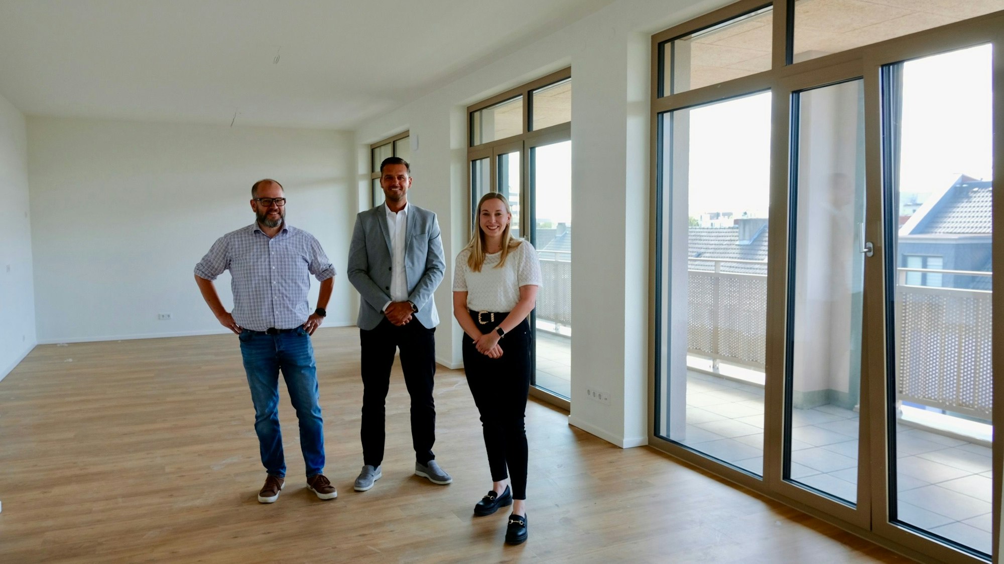 Zwei Männer und eine Frau stehen in einem leeren Raum mit großen bodentiefen Fenstern, die auf einen Balkon hinausgehen.