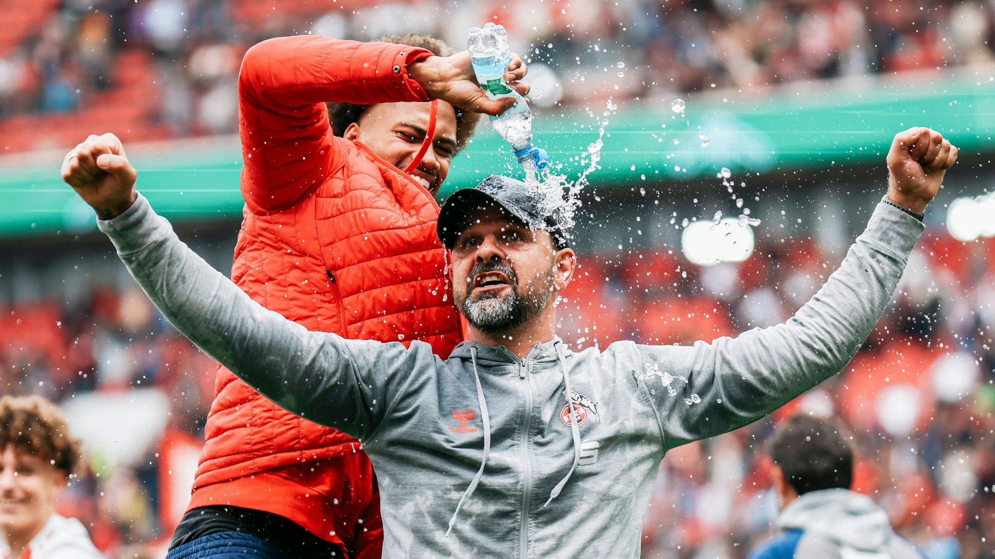 Stefan Ruthenbeck streckt jubelnd die Arme aus, während ein Spieler ihn euphorisch mit Wasser aus einer Plastikflasche überschüttet.