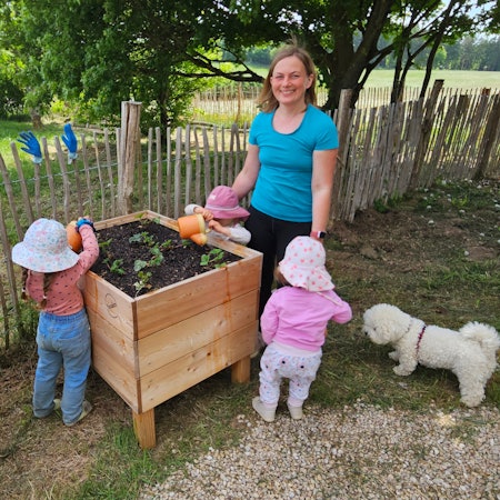 Eine Frau in türkisfarbenen T-Shirt steht mit drei kleinen Kindern und einem weißen Hund an einem Hochbeet mit Erdbeerpflanzen.