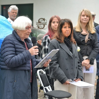Gisela Freier hält ein Mikrofon und ein Manuskript in den Händen. Daneben stehen einige der Besucher.