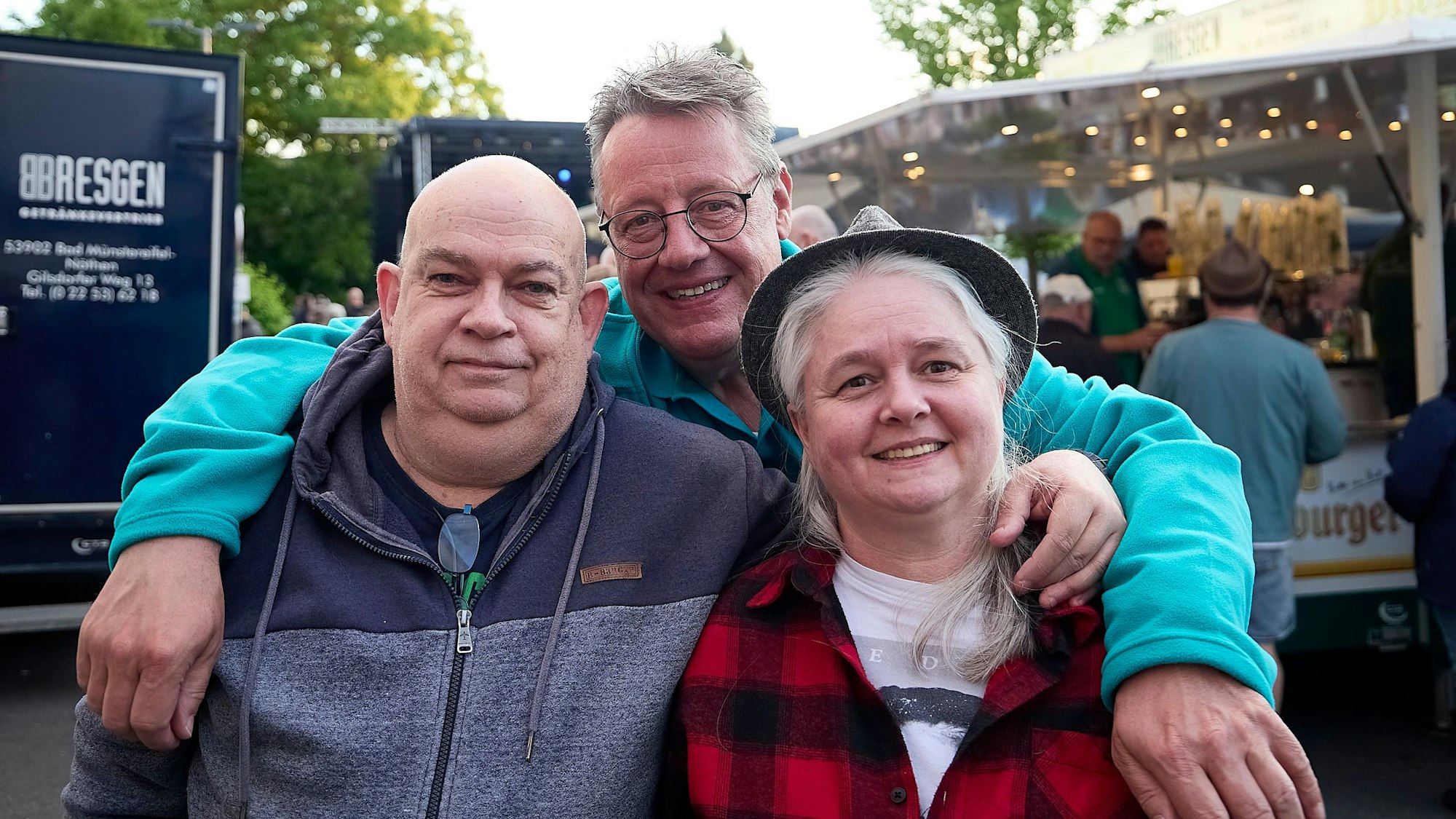 Zwei Männer und eine Frau stehen Arm in Arm vor einer Getränkebude.