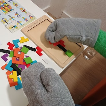 Das Bild zeigt eine Mitarbeiterin, die mit Ofenhandschuhen ein Holzpuzzle macht. Die Ofenhandschuhe simulieren eine taktile Unterempfindlichkeit und erschweren eine feinmotorische Aufgabe. Viele Menschen mit Autismus haben solche Wahrnehmungsbesonderheiten, auch wenn sie nicht zur Kernsymptomatik gehören.