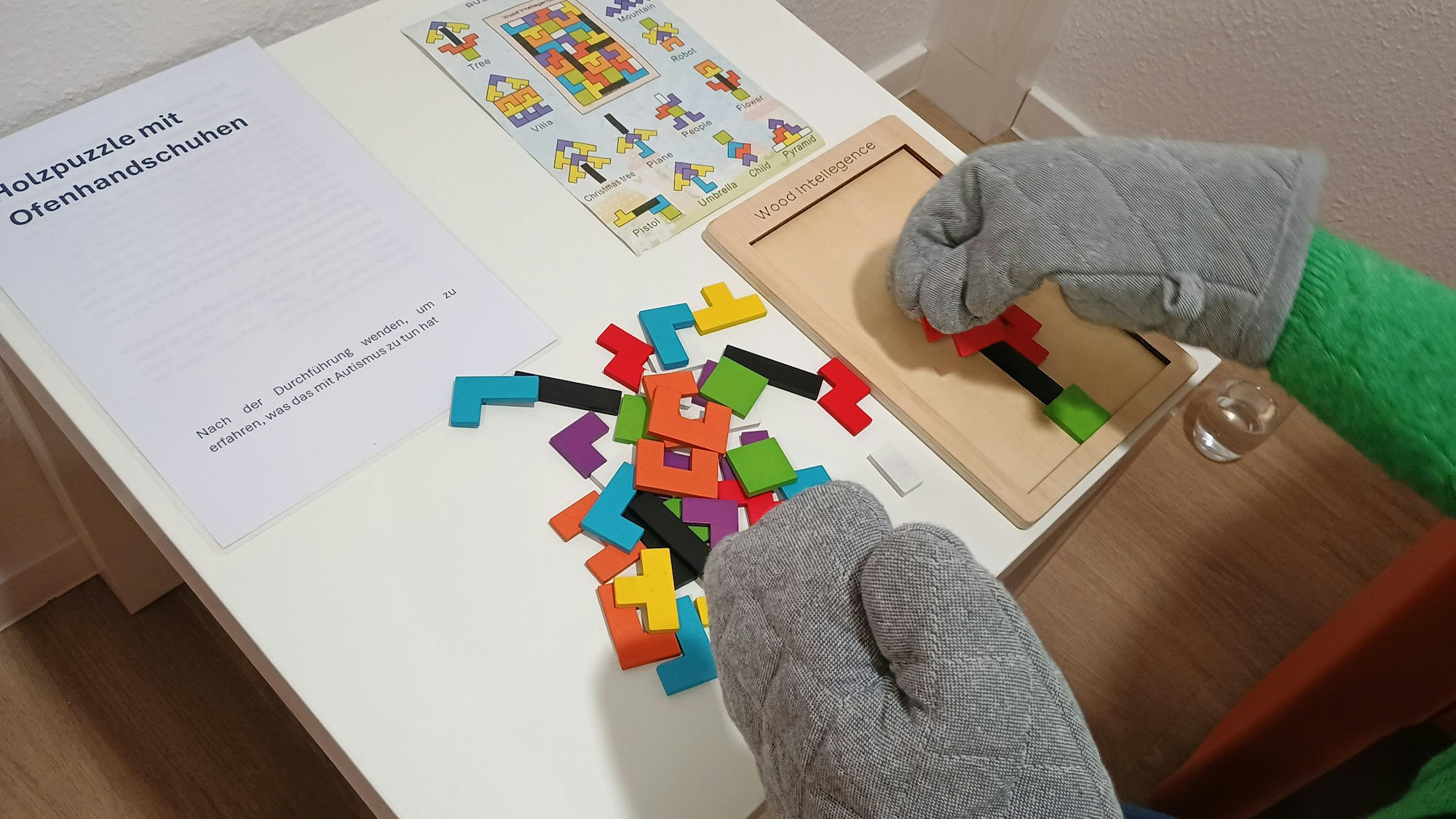 Das Bild zeigt eine Mitarbeiterin, die mit Ofenhandschuhen ein Holzpuzzle macht. Die Ofenhandschuhe simulieren eine taktile Unterempfindlichkeit und erschweren eine feinmotorische Aufgabe. Viele Menschen mit Autismus haben solche Wahrnehmungsbesonderheiten, auch wenn sie nicht zur Kernsymptomatik gehören.