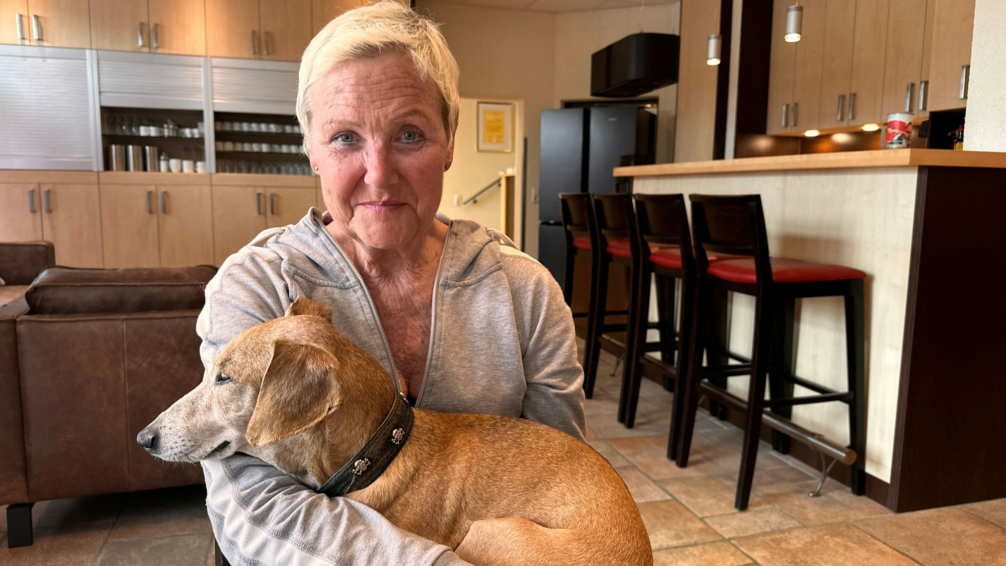 Eine Frau mit kurzen blonden Haaren sitzt an einem Tisch und trägt einen Hund auf ihrem Schoß.