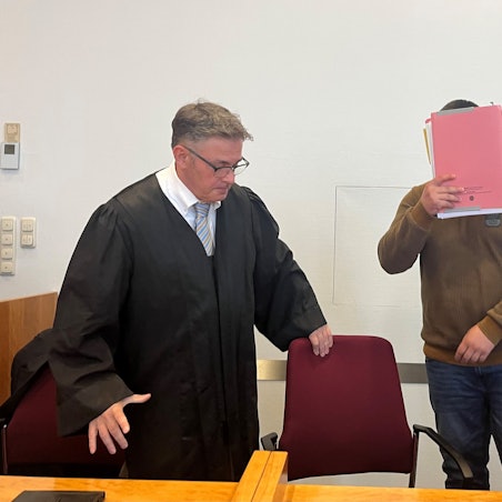 Der Angeklagte steht neben seinem Verteidiger und hält sich eine Mappe vors Gesicht.