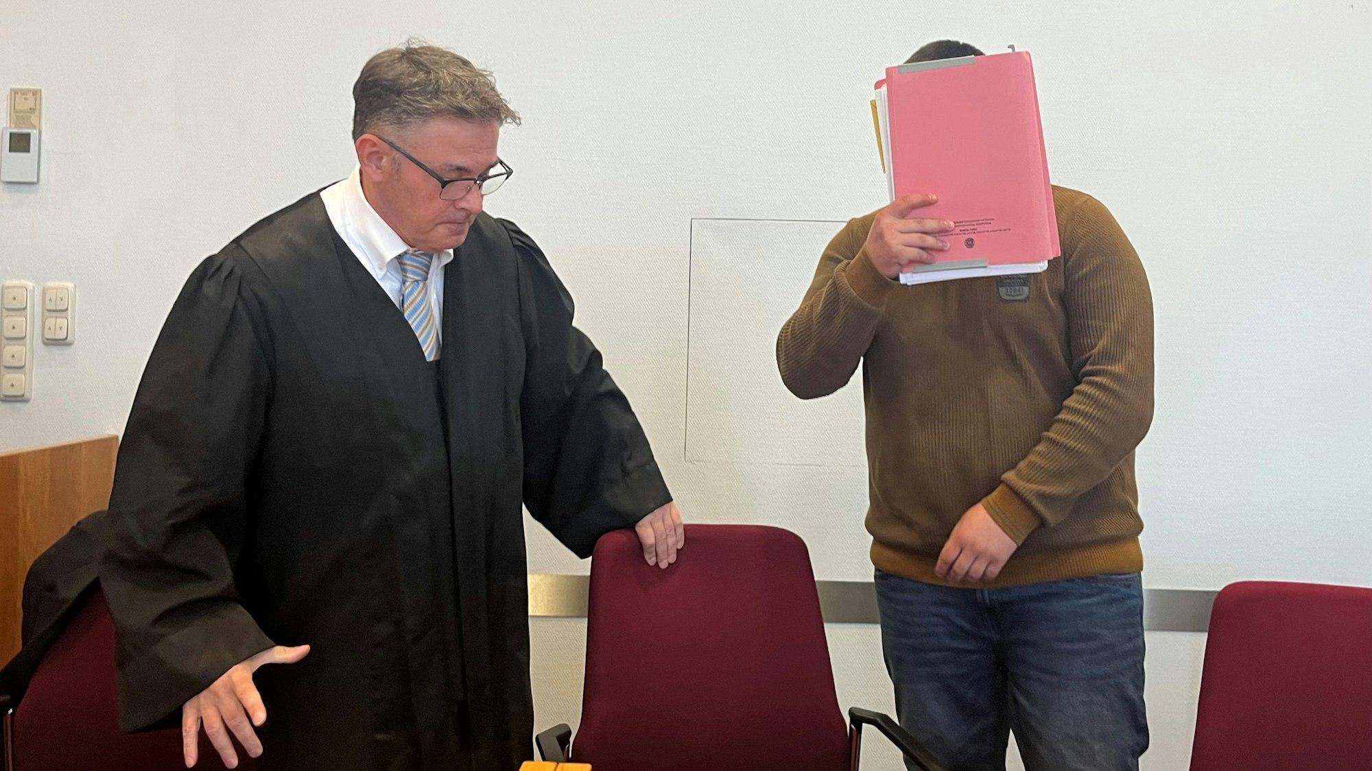 Der Angeklagte steht neben seinem Verteidiger und hält sich eine Mappe vors Gesicht.