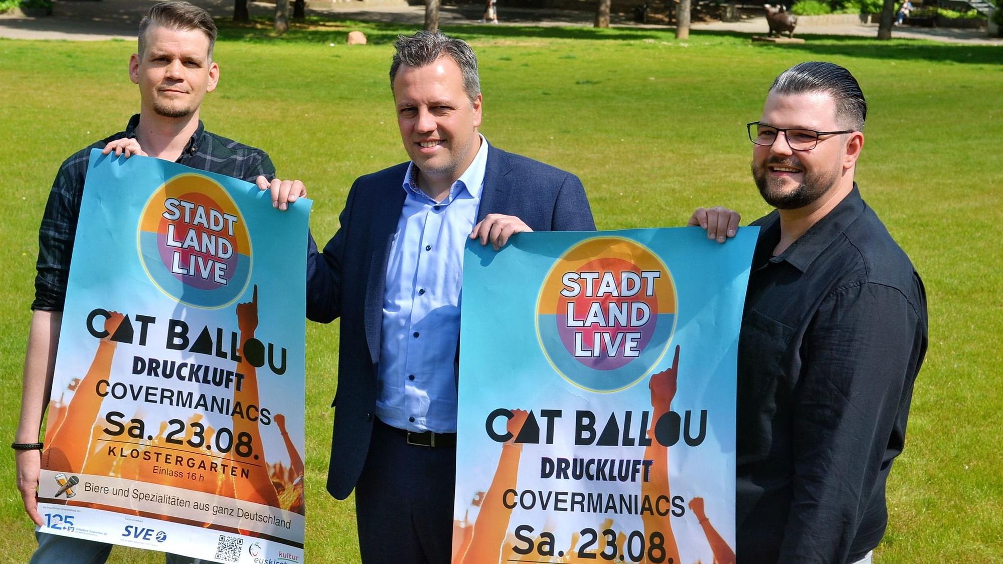 Drei Männer stehen auf einem grünen Rasen und halten zwei Plakate, mit denen ein Konzert von Cat Ballou in Euskirchen angekündigt wird.