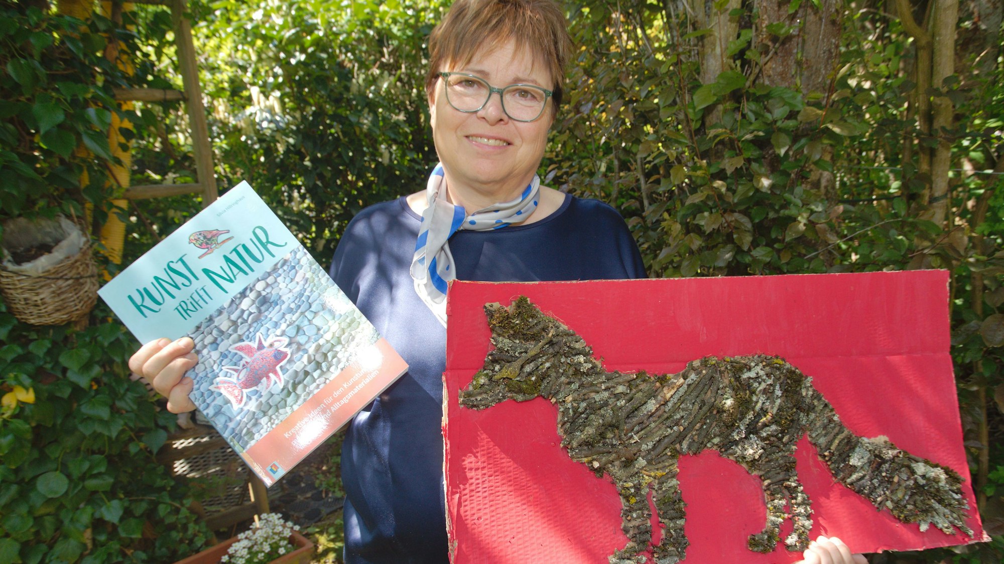 Zu sehen ist eine Frau mit einem Bild und einem Buch in den Händen.