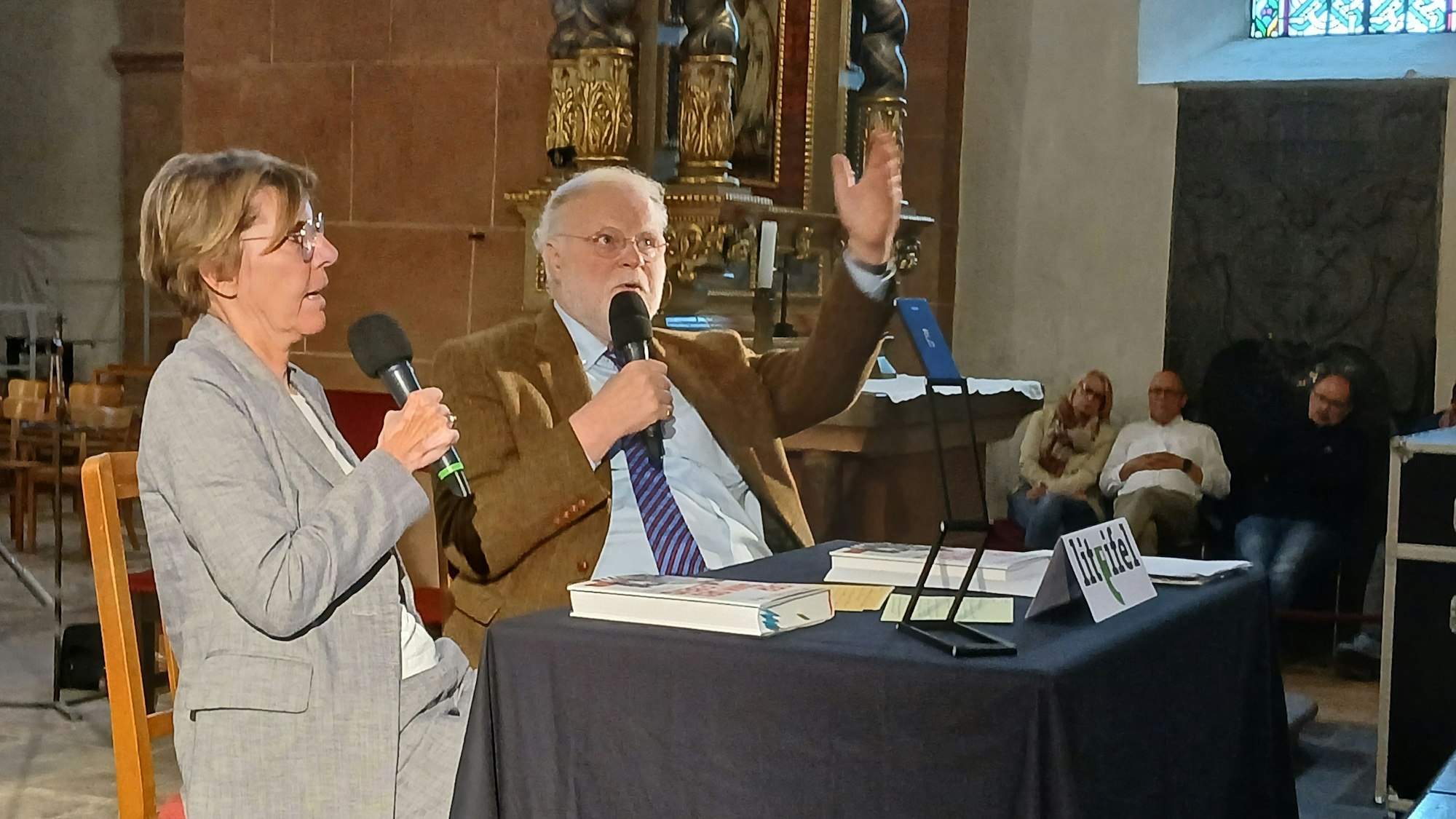 Bettina Böttinger und Dr. Manfred Lütz sitzen an einem Tisch in der Basilika in Steinfeld und sprechen in Mikrofone.