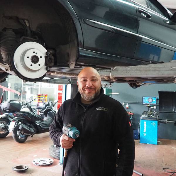Das Bild zeigt Mhedi Ghaffari in seiner Werkstatt, hinter ihm mehrere Motorräder und ein Auto auf einer Hebebühne. In der Hand hält er einen Schlagschrauber.