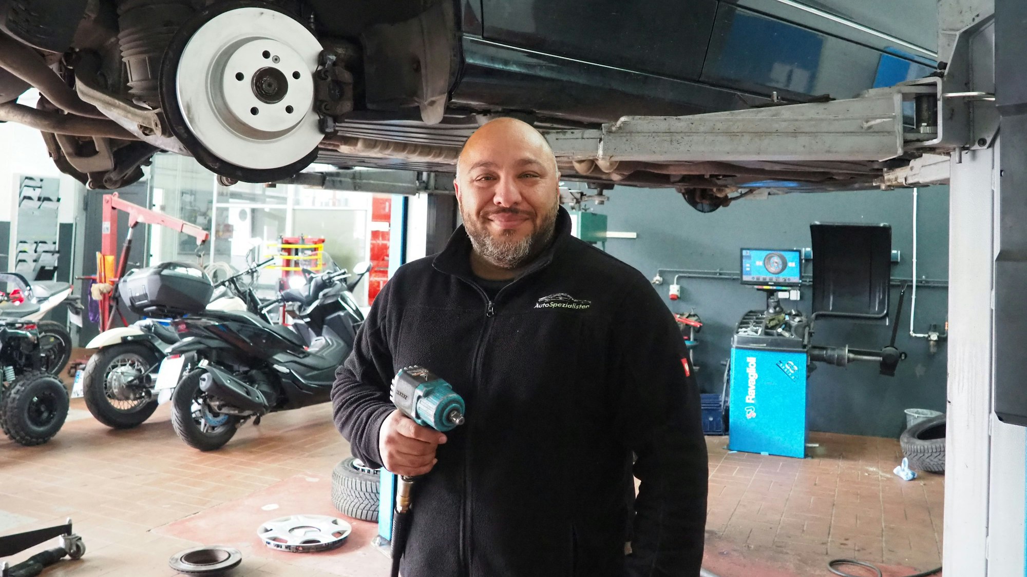 Das Bild zeigt Mhedi Ghaffari in seiner Werkstatt, hinter ihm mehrere Motorräder und ein Auto auf einer Hebebühne. In der Hand hält er einen Schlagschrauber.