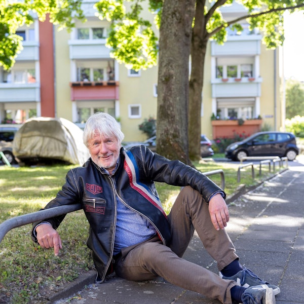 Ein Mann sitzt auf dem Bürgersteig am Rande einer von Metall-Absperrungen begrenzten Grünfläche.