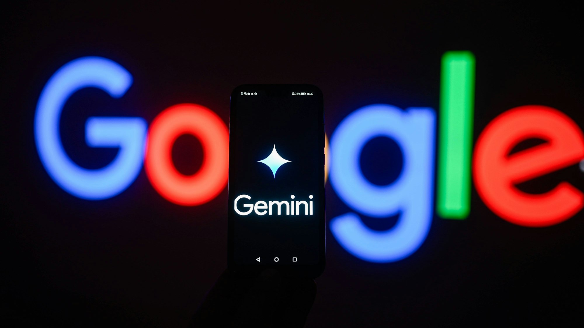 Ein Smartphone, auf dessen Bildschirm ein Gemini-Logo zu sehen ist, steht vor einem leuchtenden Google-Logo