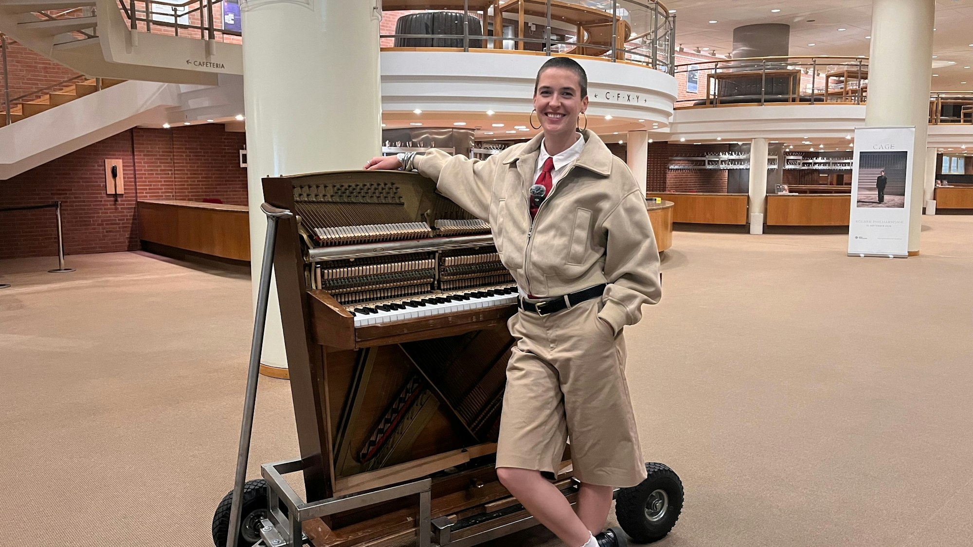 Eine junge Frau lehnt an einem Klavier mit Rollen im Foyer der Philharmonie und lächelt in die Kamera.