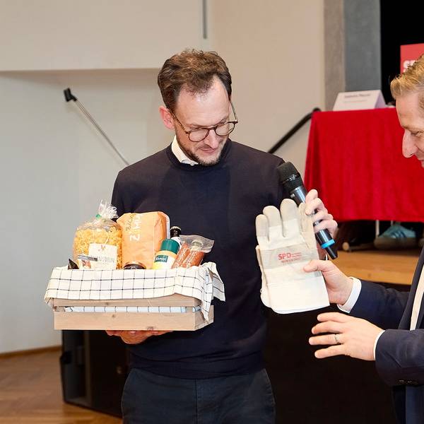 Das Bild zeigt, wie Waasem Arbeitshandschuhe mit der Aufschrift SPD an Markus Ramers übergibt. In der anderen Hand hält Waasem einen Fresskorb