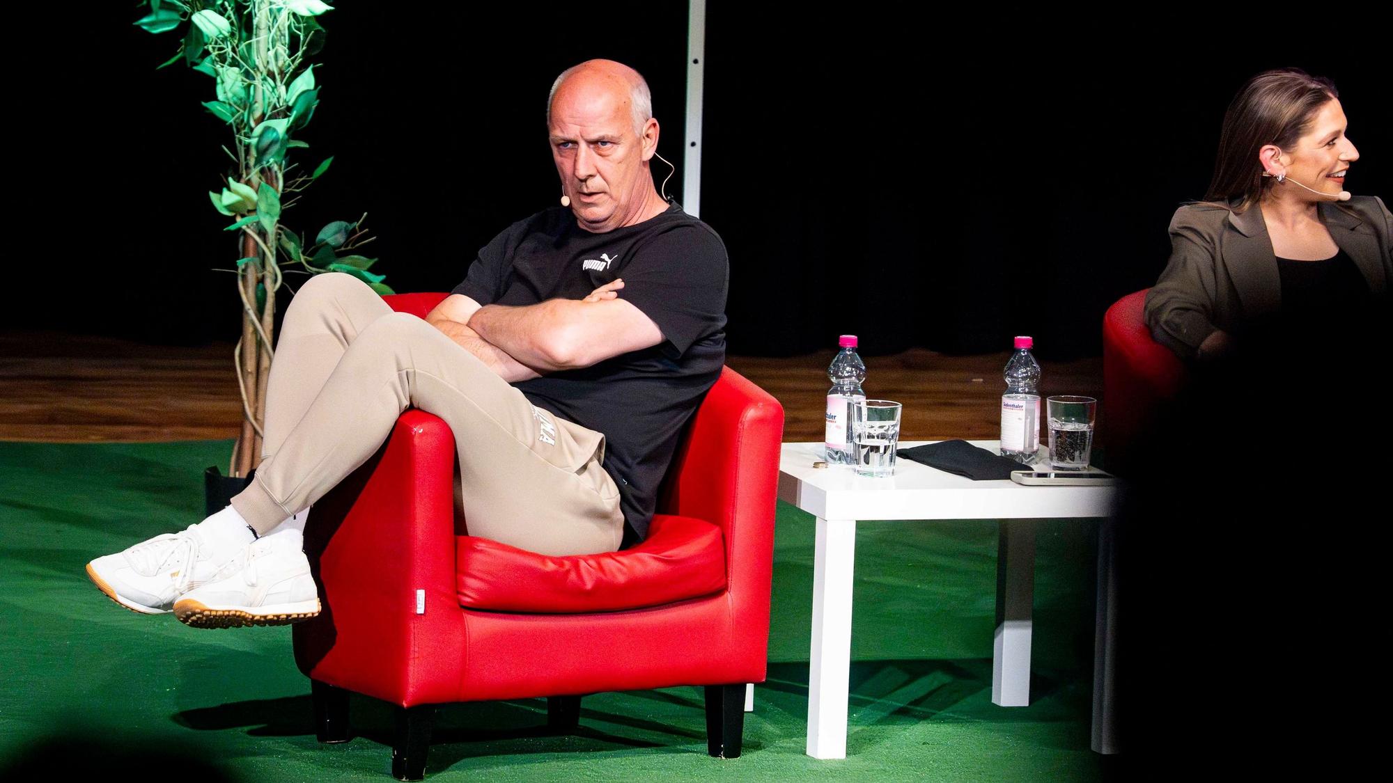 Das Bild zeigt Mario Basler, der in einem roten Sessel mit verschränkten Armen sitzt.