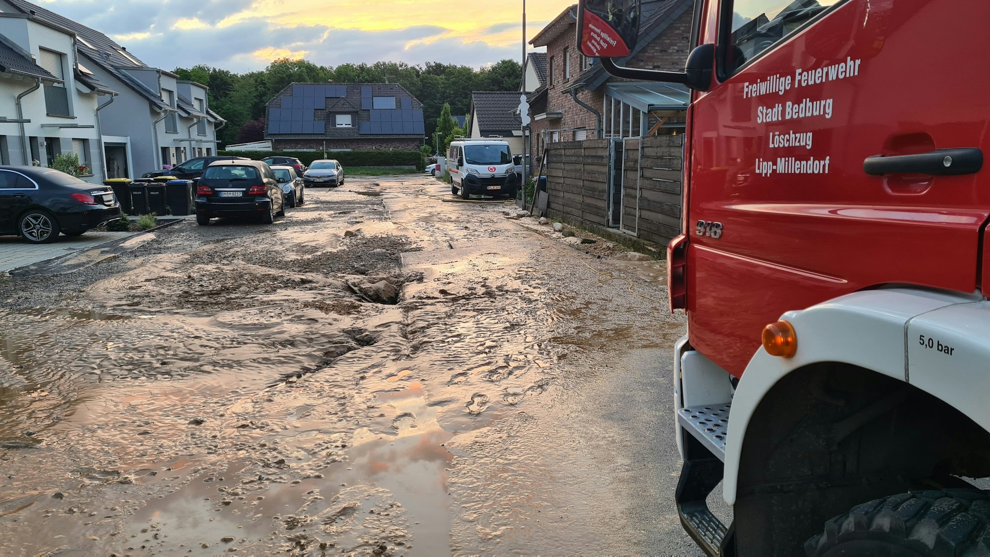 Das Foto zeigt eine verschlammte Straße, an der Seite steht ein Einsatzfahrzeug der Feuerwehr.