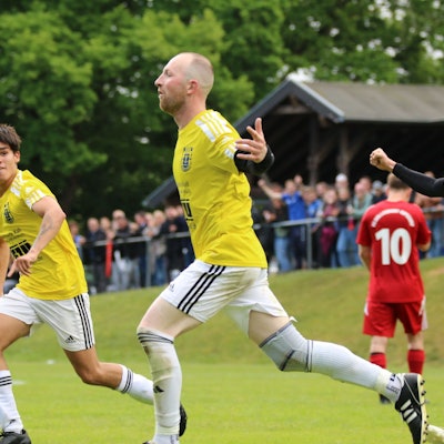 Ein Fußballer läuft mit ausgestreckten Armen nach einem Treffer über den Platz. Auch seine Mitspieer jubeln.