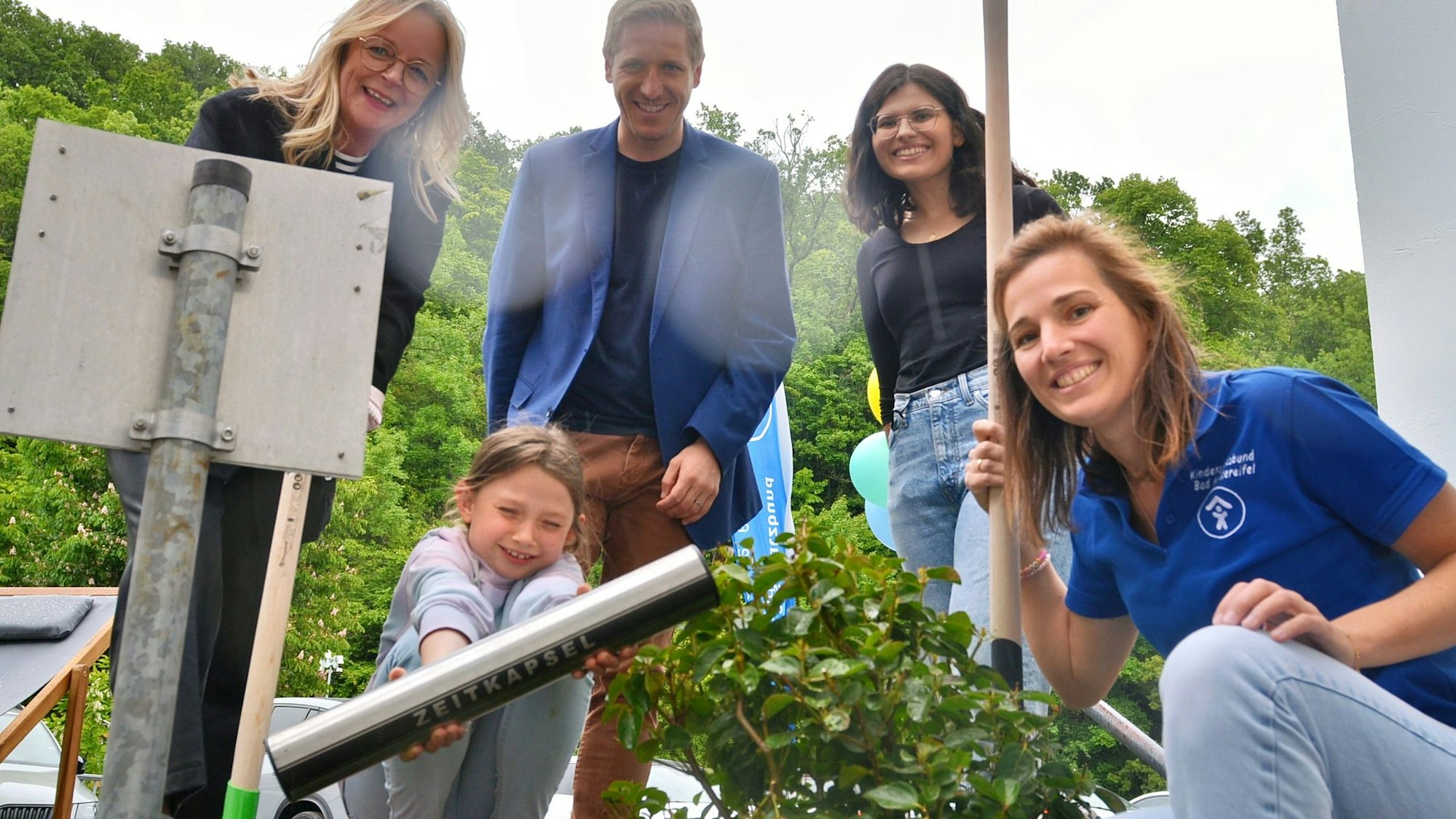 Ein Mädchen hält die Zeitkapsel in der Hand, die eingegraben wird. Erwachsene stehen um sie herum.