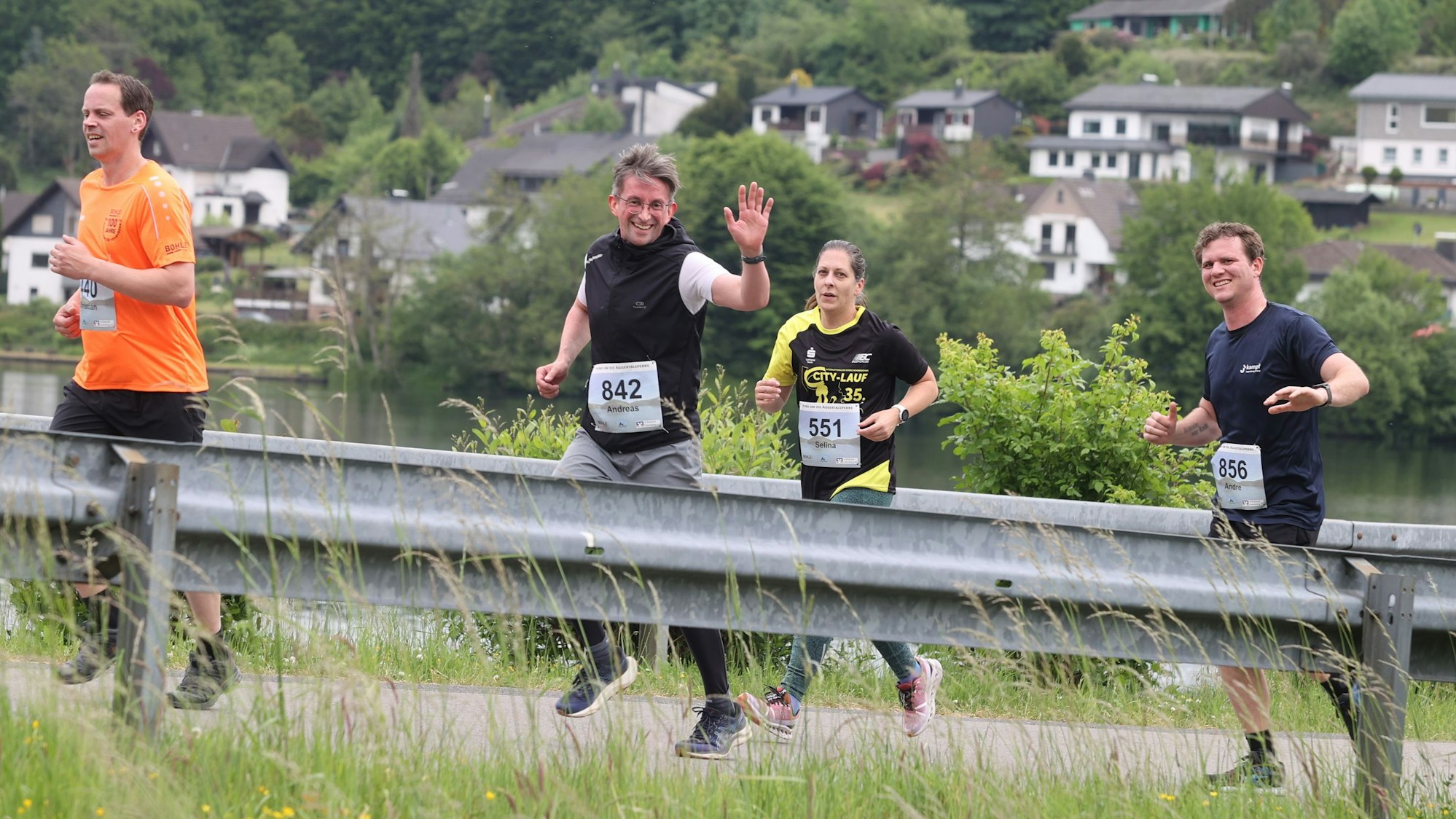 Blick auf drei Männer und eine Frau, die an der Aggertalsperre entlanglaufen. Einer der Männer winkt.
