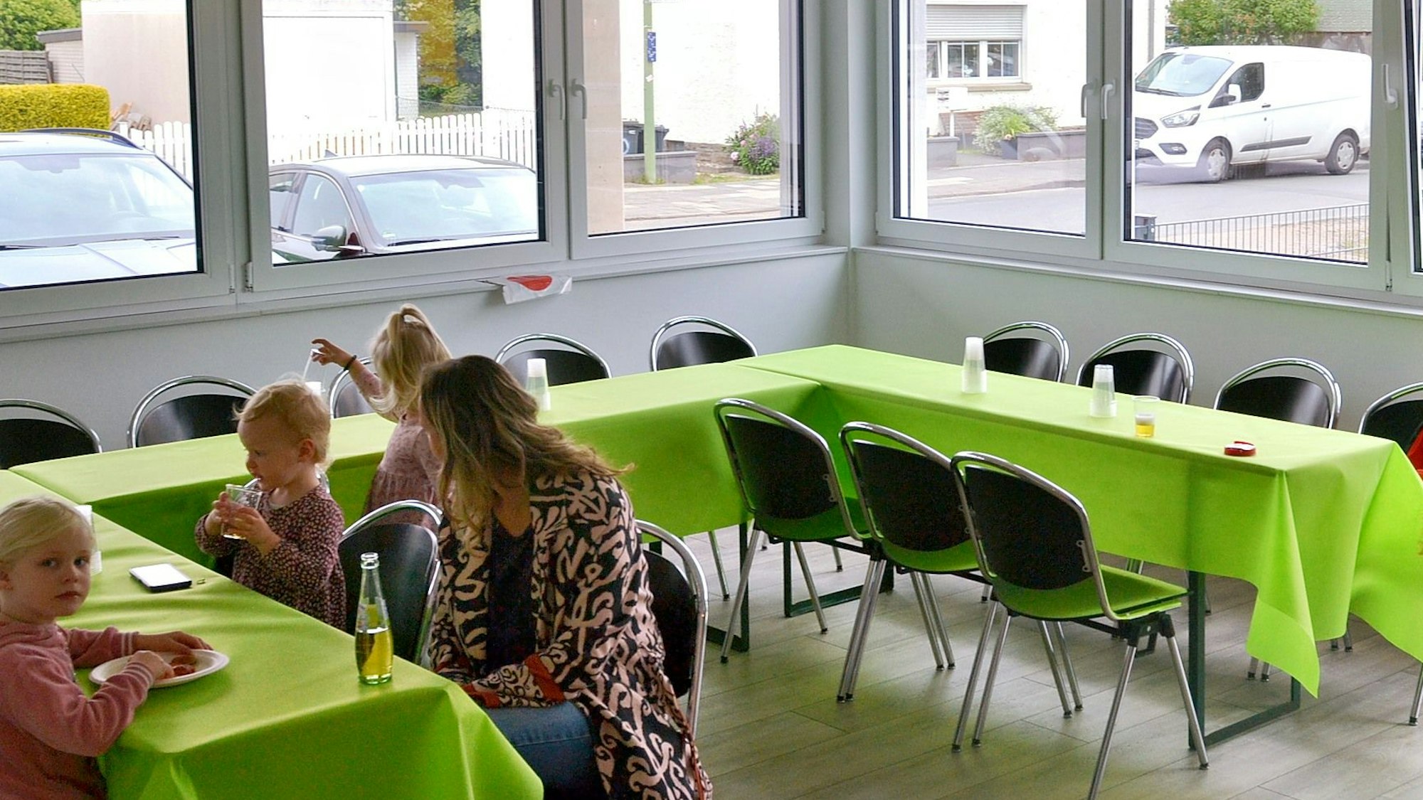 In einem großen Raum stehen aneinander gereihte Tische, auf denen grüne Tischdecke liegen. Eine Frau und ein paar Kinder sitzen daran.