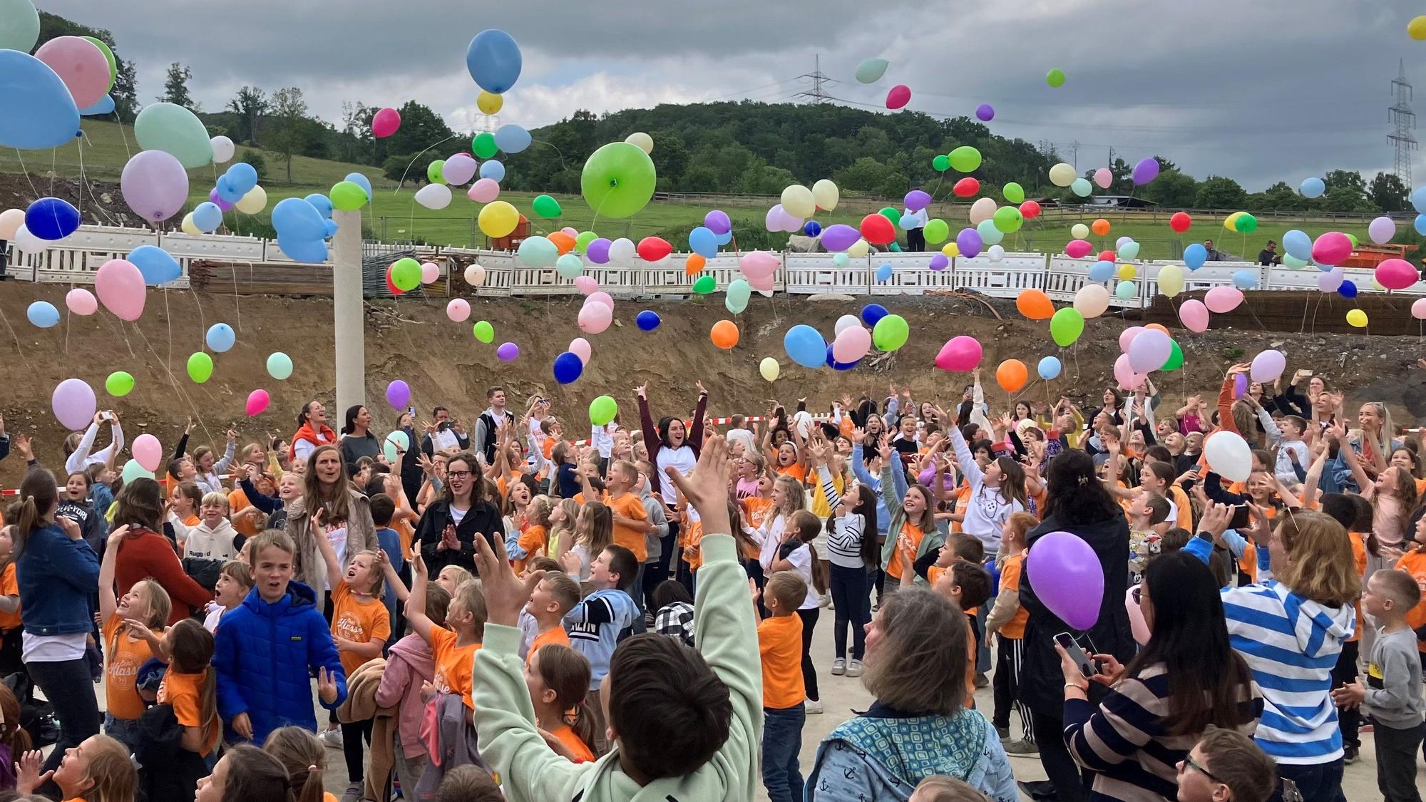 Schülerinnen und Schüler sowie Lehrkräfte lassen bunte Luftballons steigen.