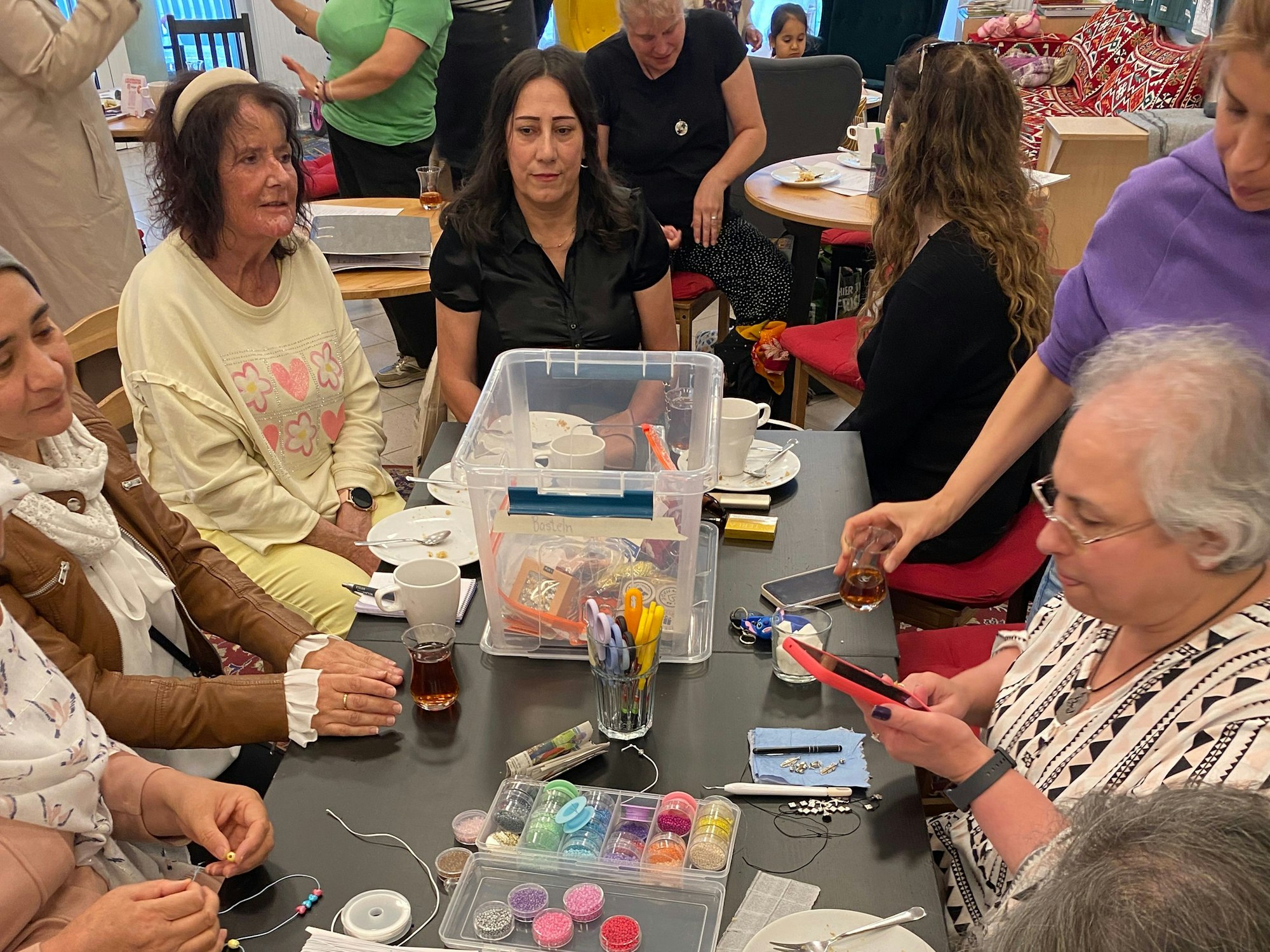 Viele Frauen sitzen an einem Tisch, auf dem Perlen in allen Größen und Farben liegen, mit denen sie Ketten und Armbänder gestalten.