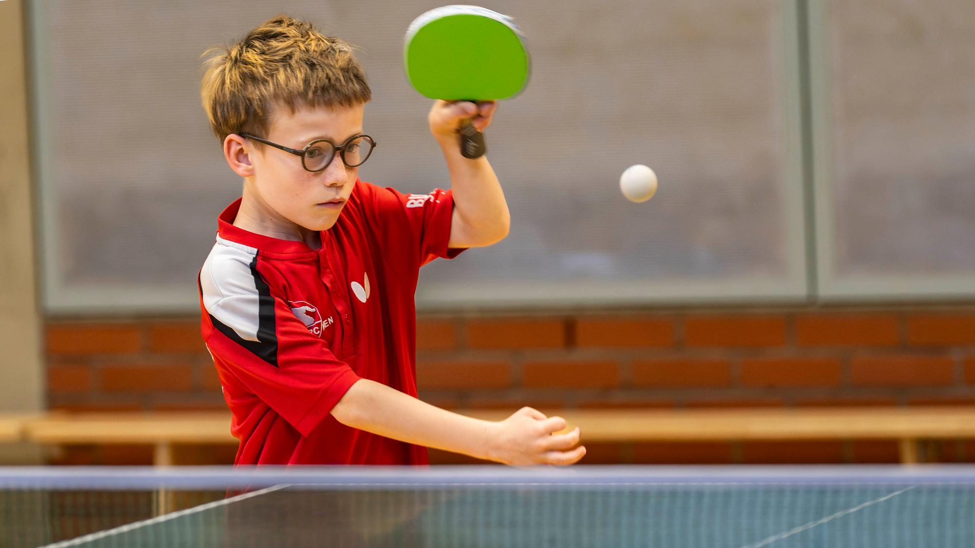 Ein Junge in einem roten Trikot und mit Brille schlägt einen Tischtennisball.