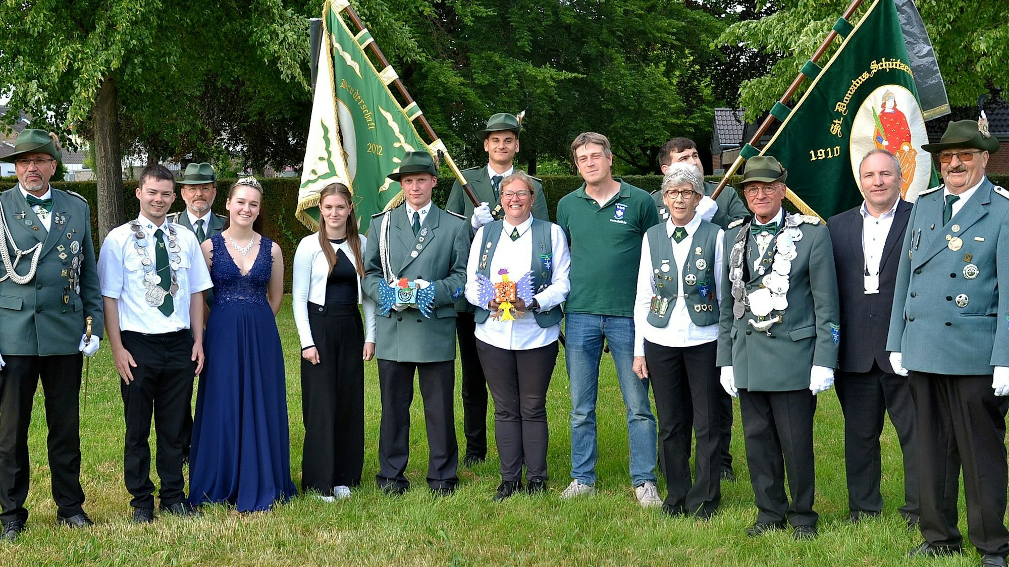 Eine Gruppe Menschen steht auf einer Wiese, manche tragen eine grün Uniform, im Hintergrund sind zwei Fahnen zu erkennen.