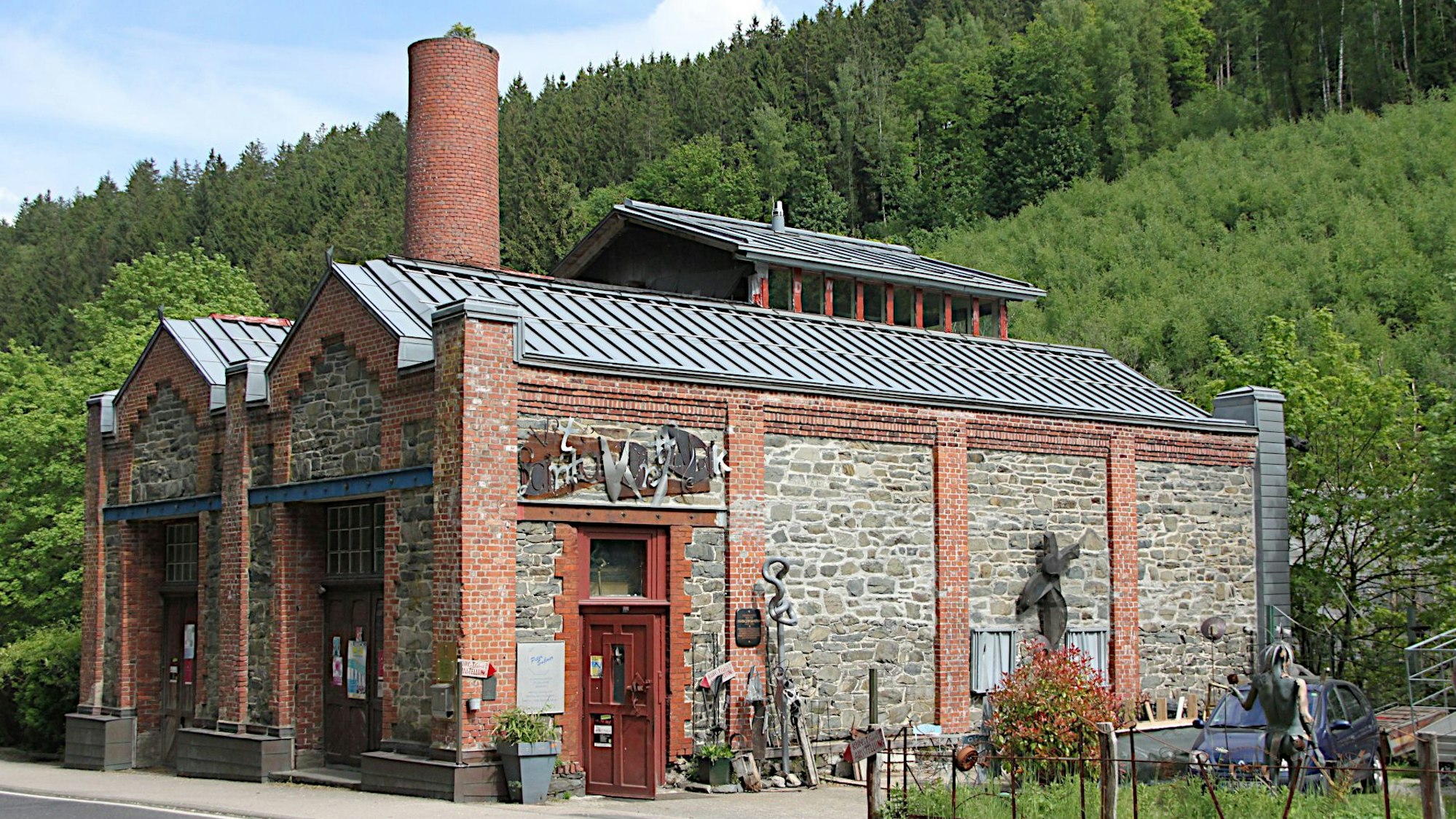 Ein altes Backsteingebäude mit großem Schornstein steht vor einem bewaldeten Hügel.