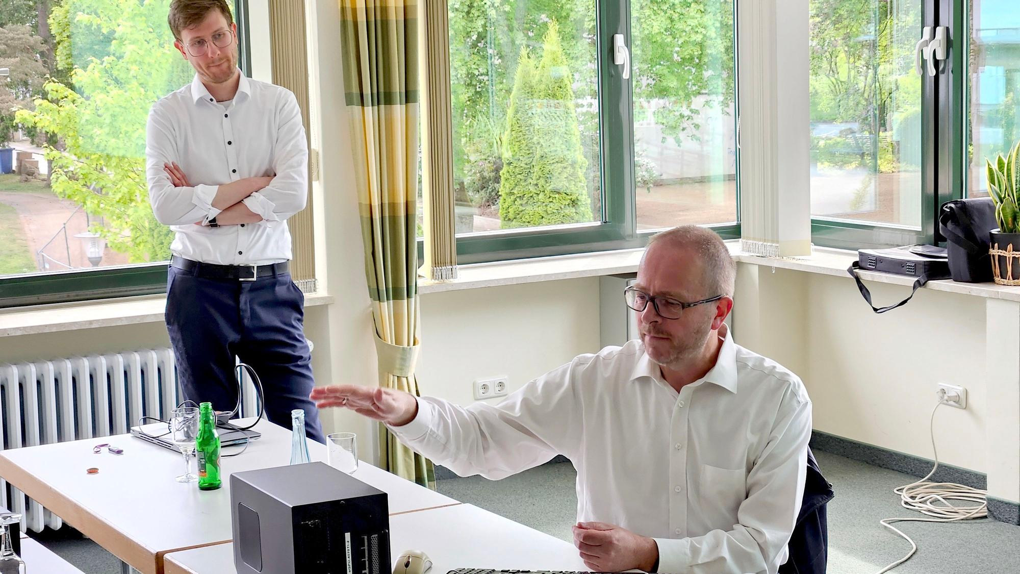 Ein Mann in weißem Hemd sitzt an einem Tisch und deutet auf eine schwarze Kiste, die auf dem Tisch steht. Ein anderer Mann steht daneben und schaut zu.