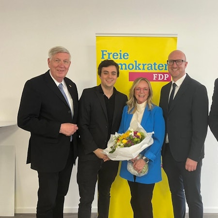 Die Herren haben die Kandidatin, die einen Blumenstrauß in den Händen hält, in ihre Mitte genommen. Dahinter steht ein Aufsteller mit der Aufschrift Freie Demokraten FDP.