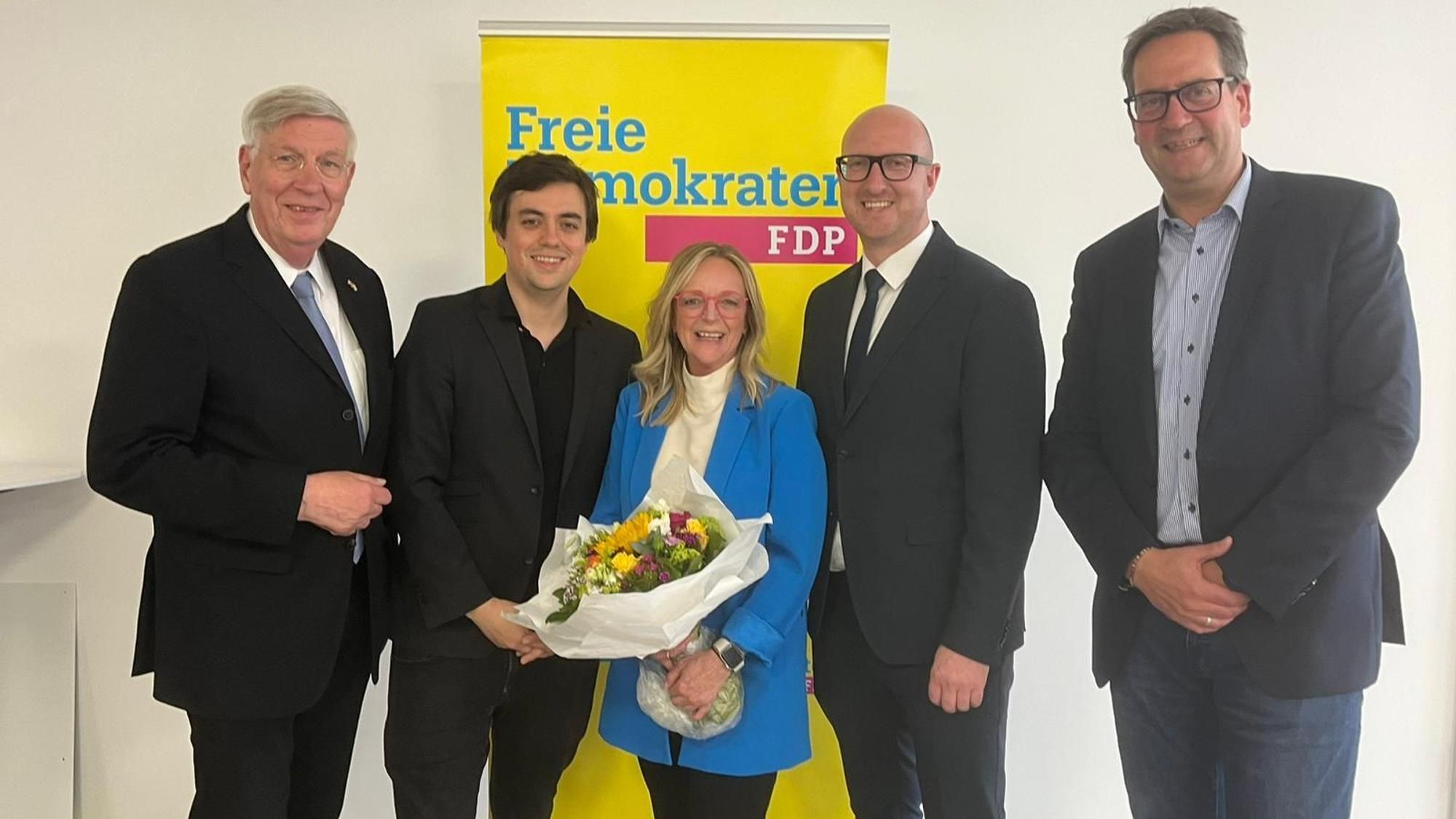 Die Herren haben die Kandidatin, die einen Blumenstrauß in den Händen hält, in ihre Mitte genommen. Dahinter steht ein Aufsteller mit der Aufschrift Freie Demokraten FDP.