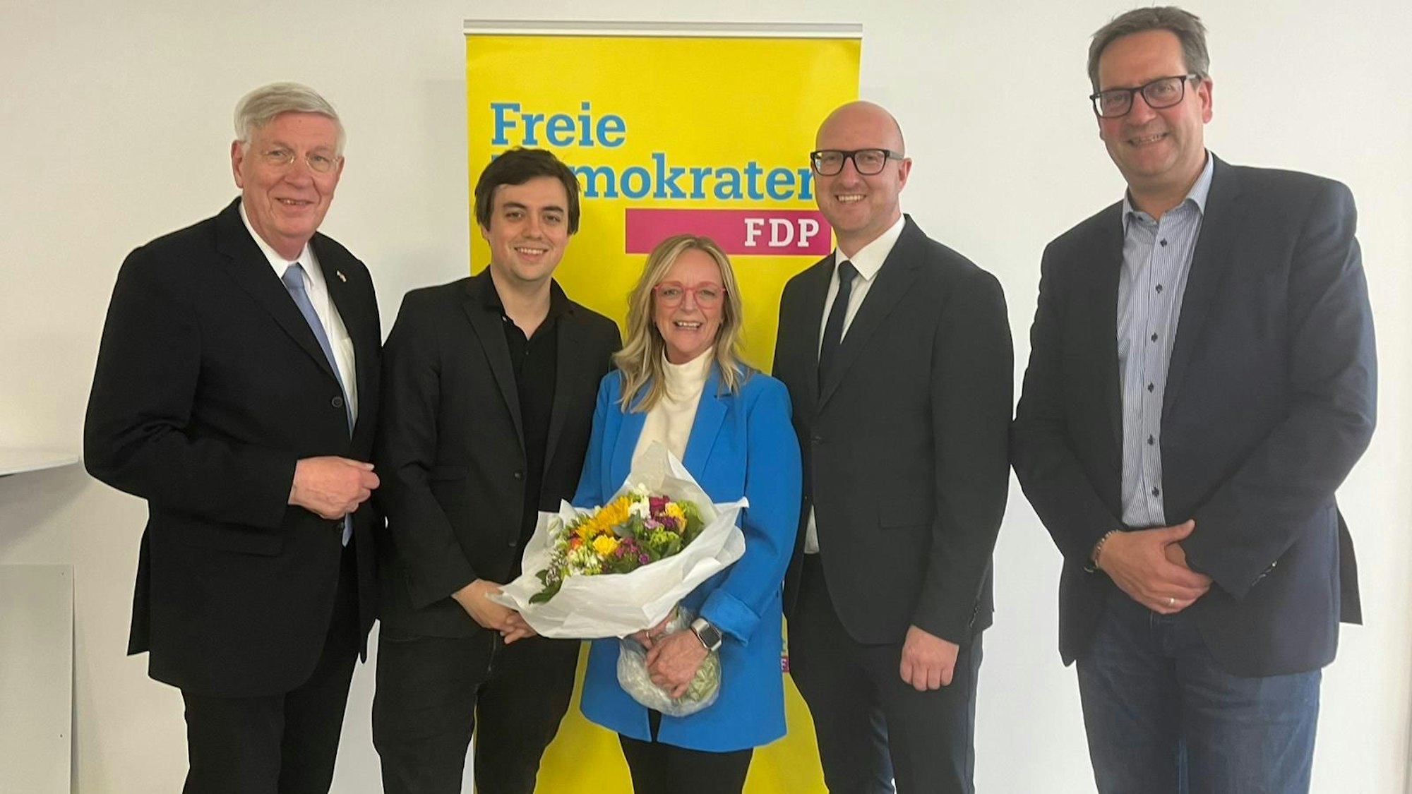 Die Herren haben die Kandidatin, die einen Blumenstrauß in den Händen hält, in ihre Mitte genommen. Dahinter steht ein Aufsteller mit der Aufschrift Freie Demokraten FDP.