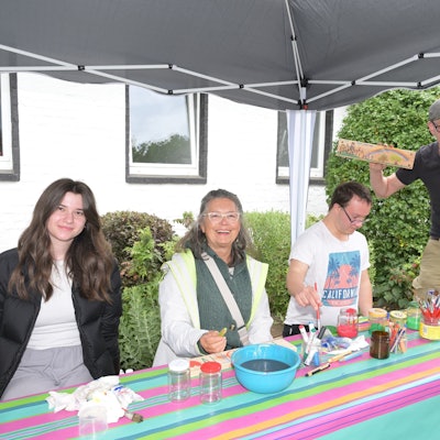 Drei Menschen sitzen vor einem Tisch und bemalen Schilder, einer steht daneben und hält die Schilder hoch.
