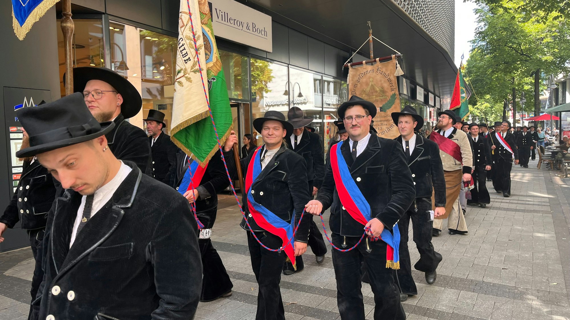 Man sieht mehrere Männer in einer Reihe in schwarzen Anzügen. Manche halten Fahnen in der Hand.