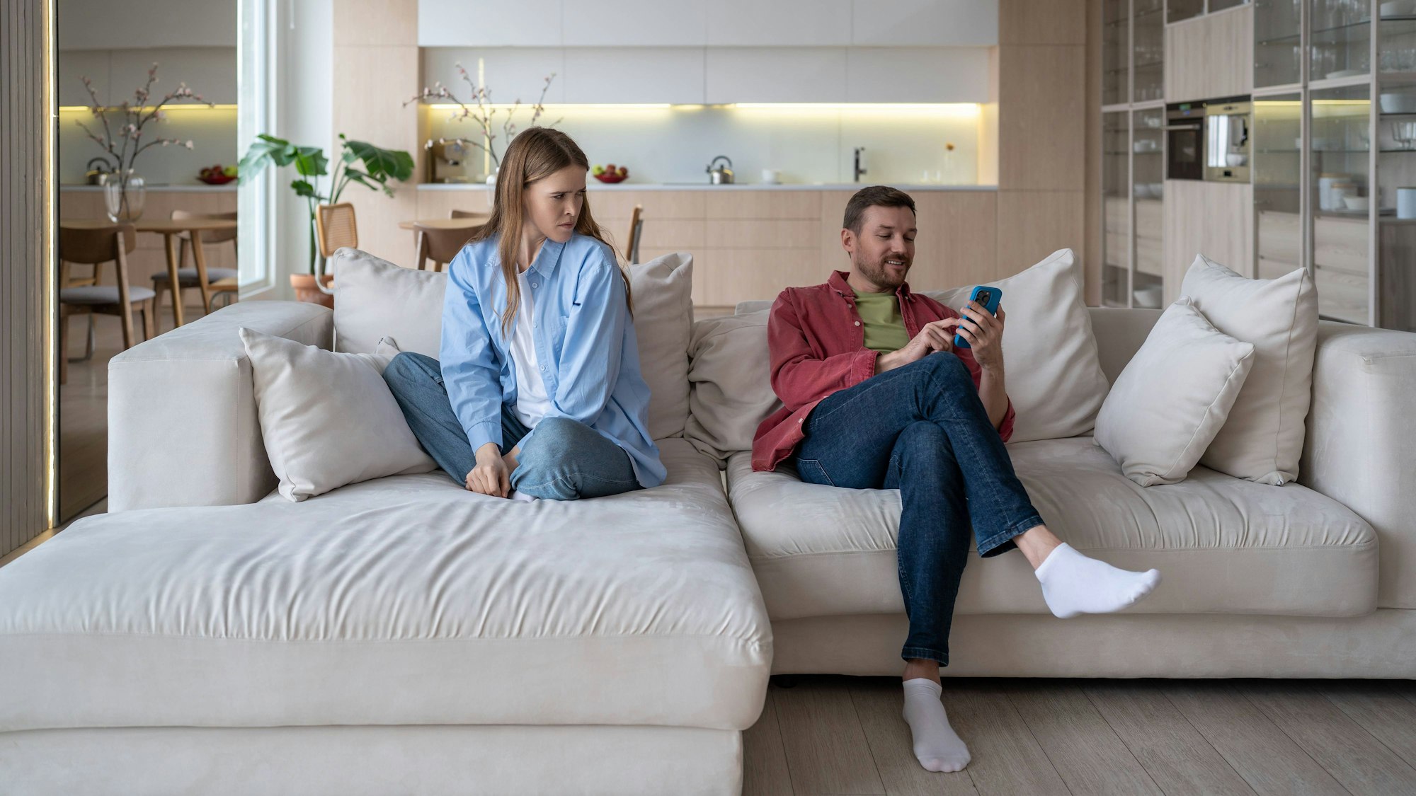 Ein Mann schaut süffisant lächelnd auf sein Handy, während eine Frau ihn enttäuscht und traurig anschaut. Beide sitzen auf einem Sofa.
