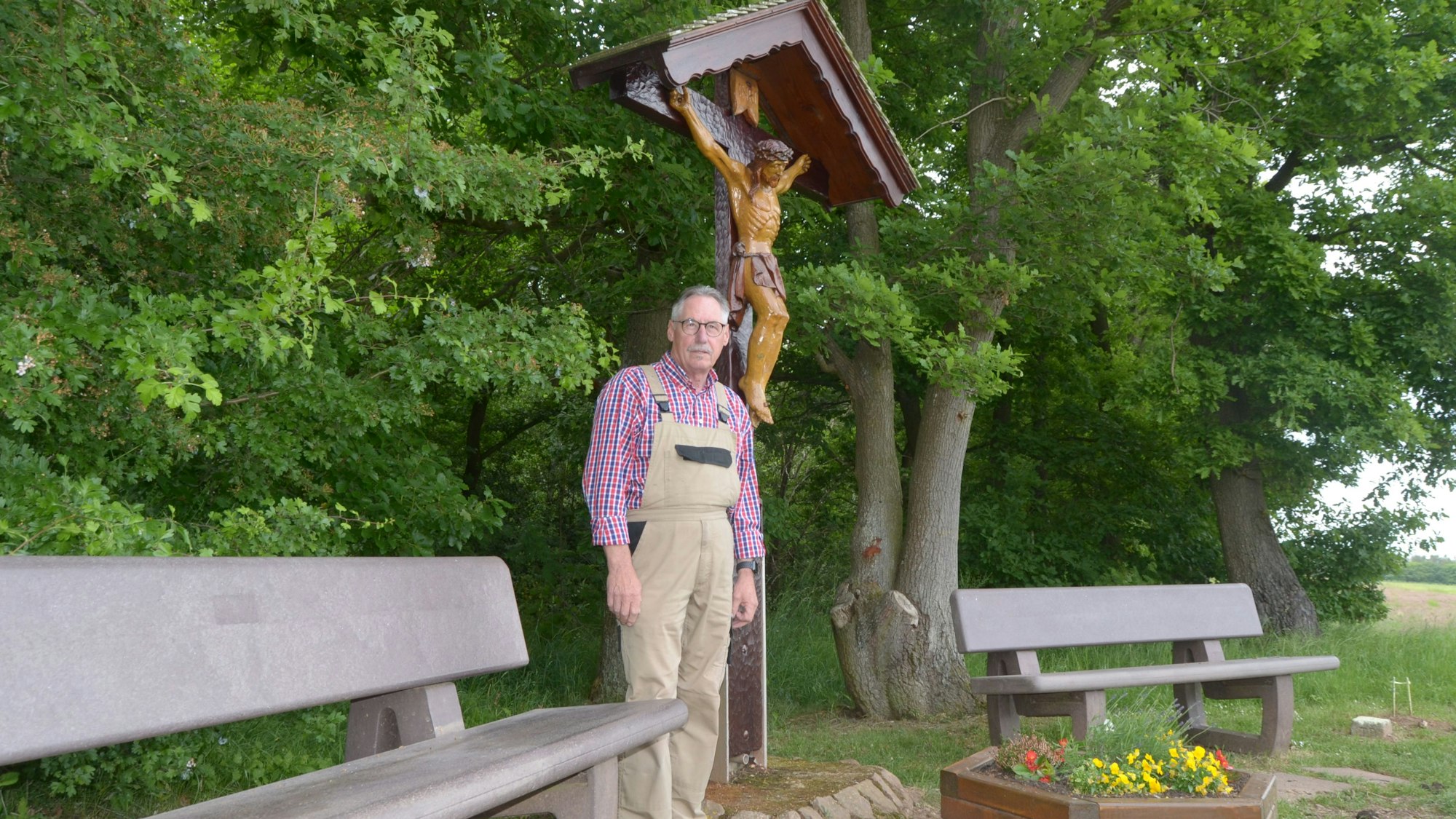 Engelbert Kolvenbach steht an einem kleinen Platz mit einem Wegekreuz und zwei Bänken bei Euskirchen, im Hintergrund sind Bäume zu sehen.