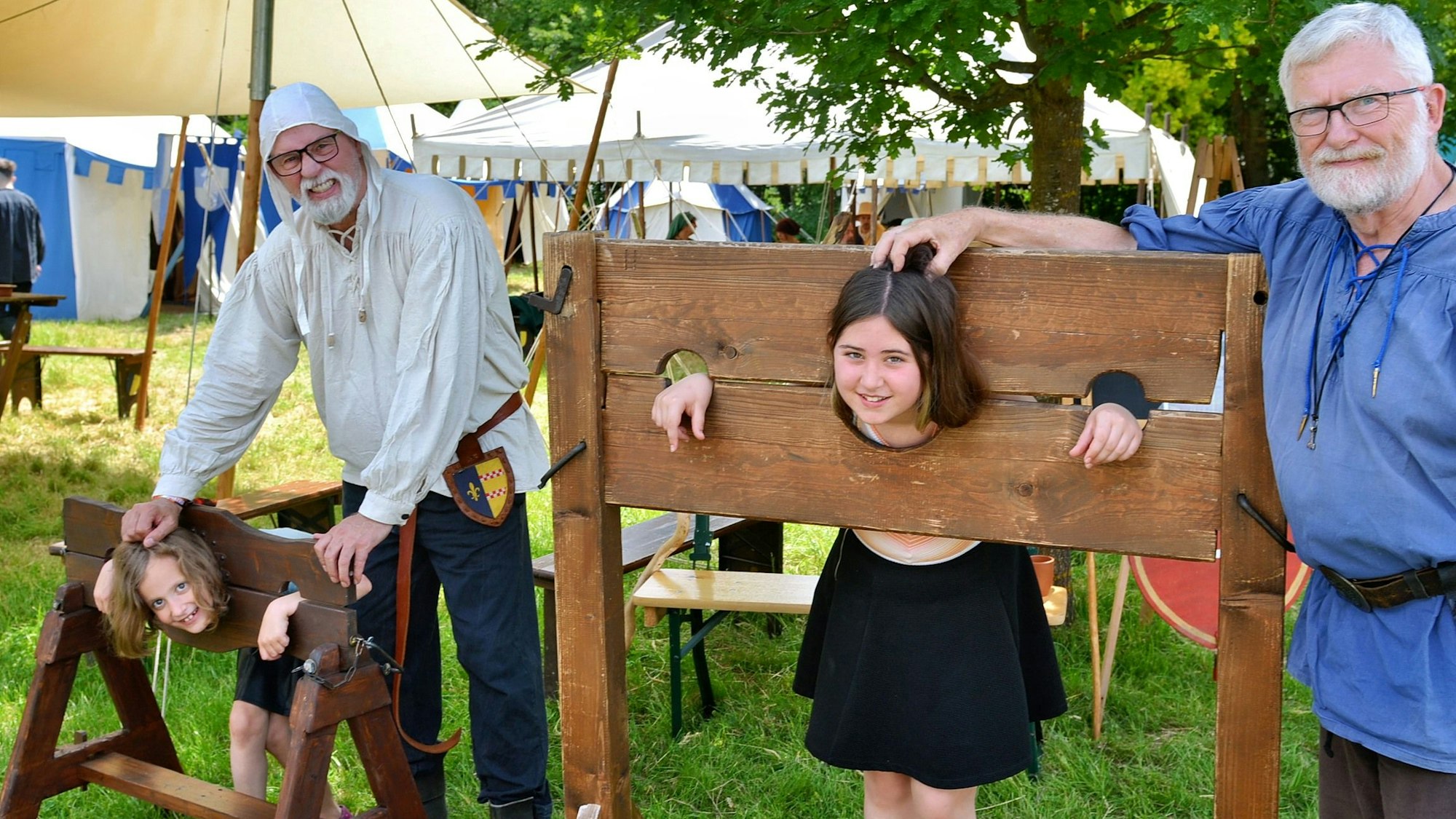 Zwei Kinder stehen jeweils in einem Pranger, daneben stehen zwei Männer in Gewändern der Ritterzeit.