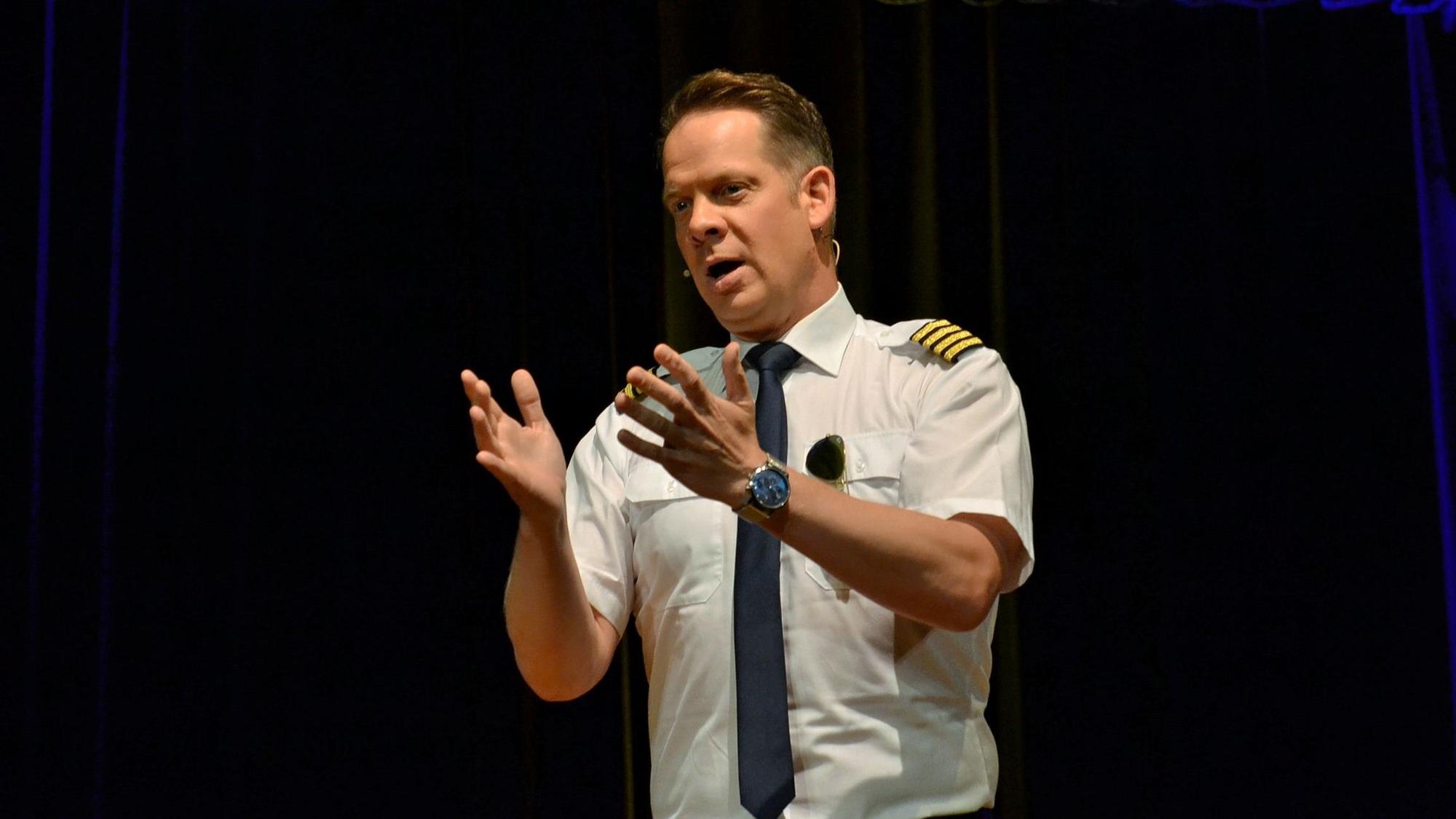 Als Pilot verkleidet, gestikuliert Marco Brüser mit beiden Händen in Richtung Publikum.