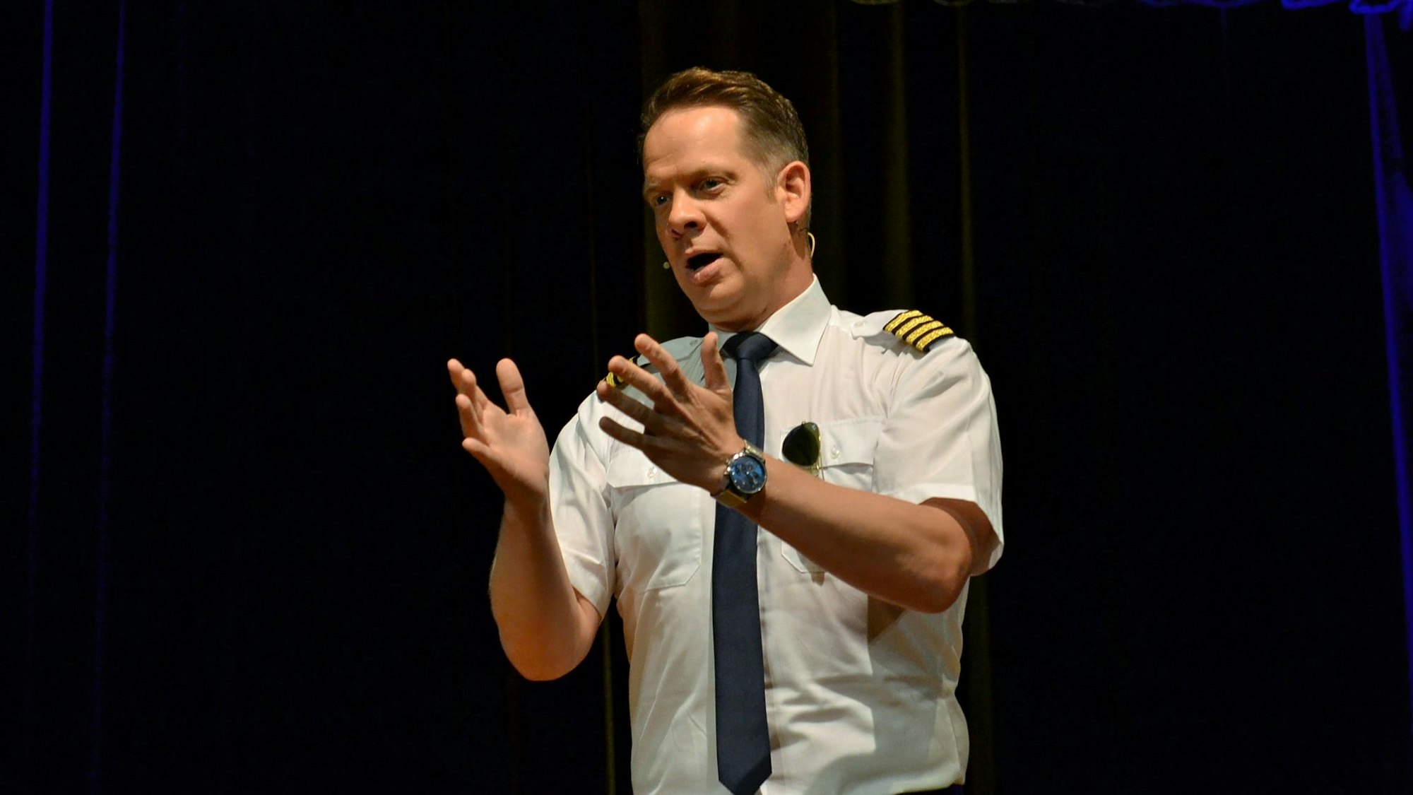 Als Pilot verkleidet, gestikuliert Marco Brüser mit beiden Händen in Richtung Publikum.
