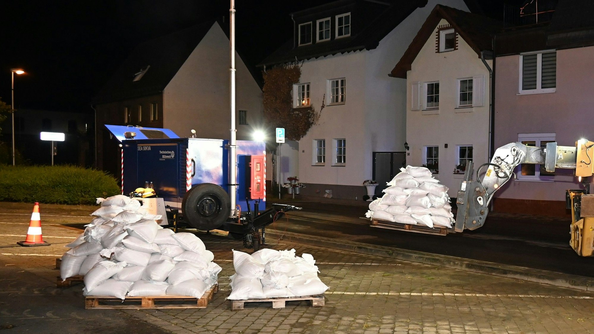 Das Bild zeigt einen Bagger, der Paletten mit Sandsäcken auf einem Parkstreifen abstellt.