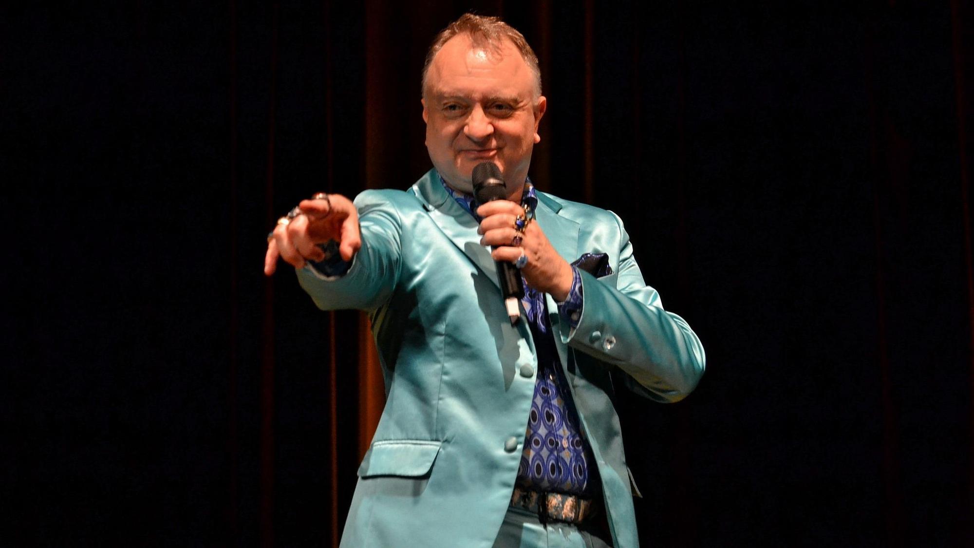 In einem blau-grünen Anzug steht Horst Blue auf der Bühne und zeigt mit dem Zeigefinger der linken Hand ins Publikum.