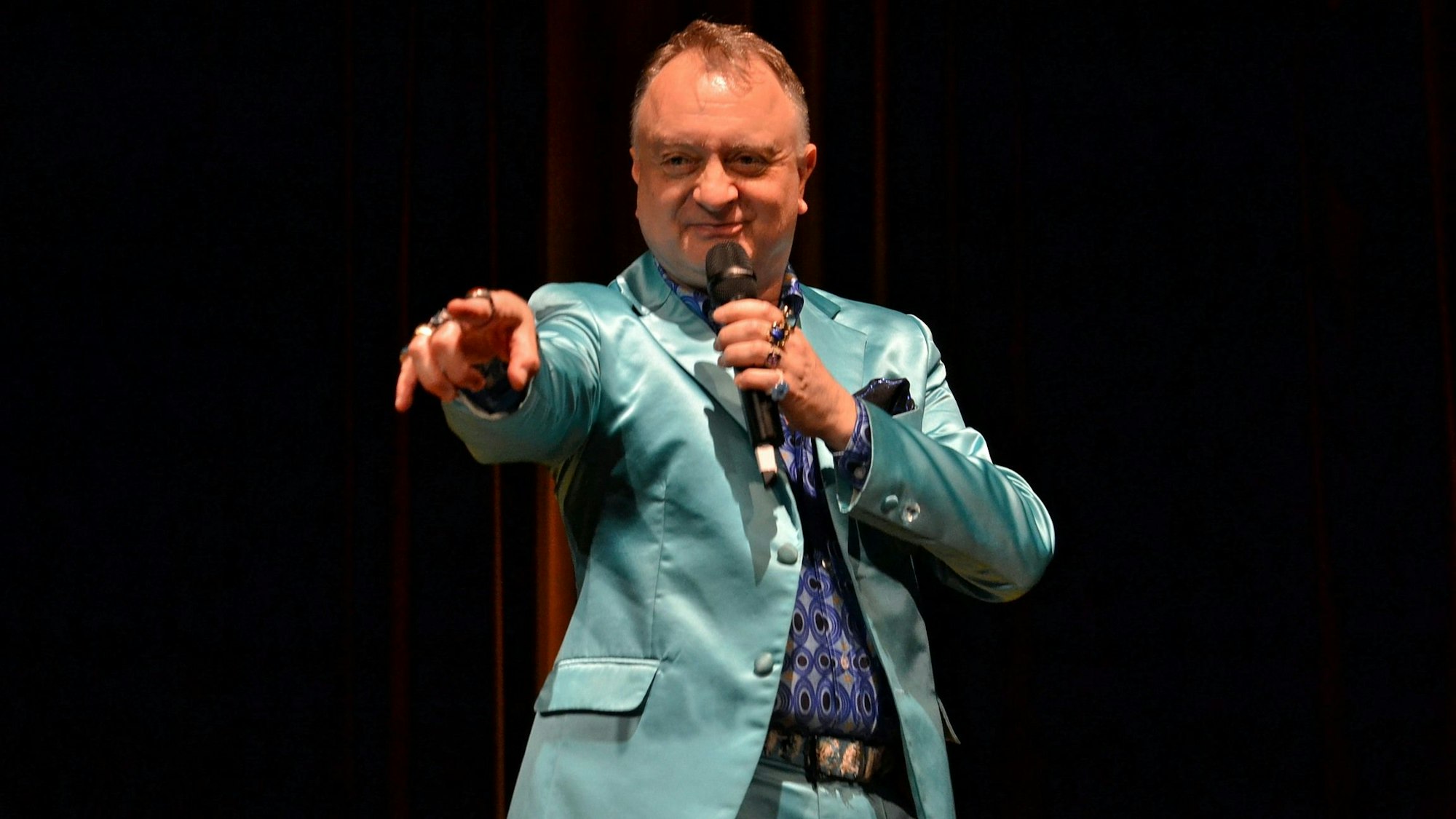 In einem blau-grünen Anzug steht Horst Blue auf der Bühne und zeigt mit dem Zeigefinger der linken Hand ins Publikum.