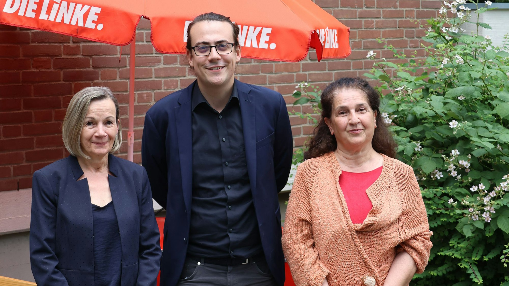 Die drei Kandidaten stehen nebeinander, hinter ihnen ein roter Sonnenschirm mit der Aufschrift „Die Linke“
